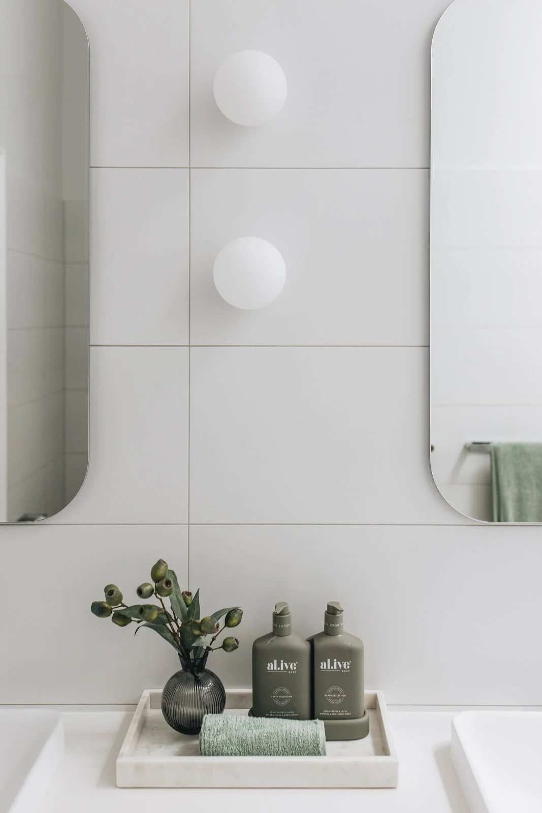 Bathroom mirror with white tiles, two spherical light fixtures above, a small vase with greenery, two bottles of lotion, a rolled green towel, and a green towel hanging in the background.