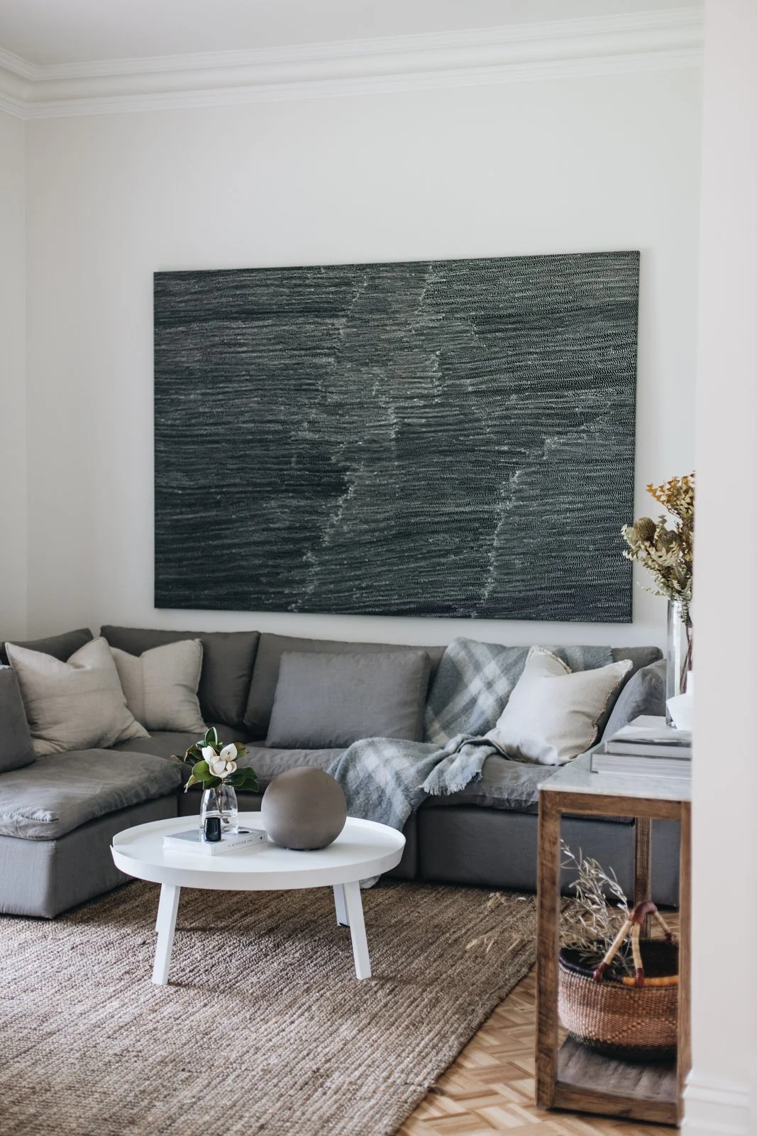Living room with a gray sectional sofa, pillows, a white coffee table with a vase and a sphere, abstract black wall art, and a wooden side table with a plant in a basket.