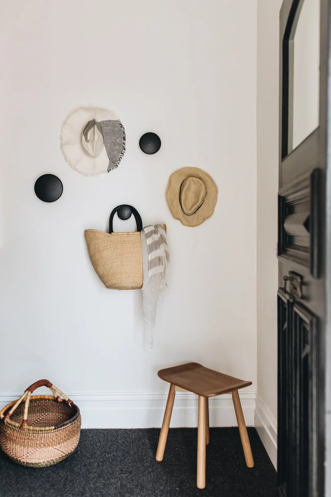 Entryway wall decor with hanging hats, a woven basket bag, and circular wall hooks, next to a small wooden stool and a woven basket on a dark carpet.