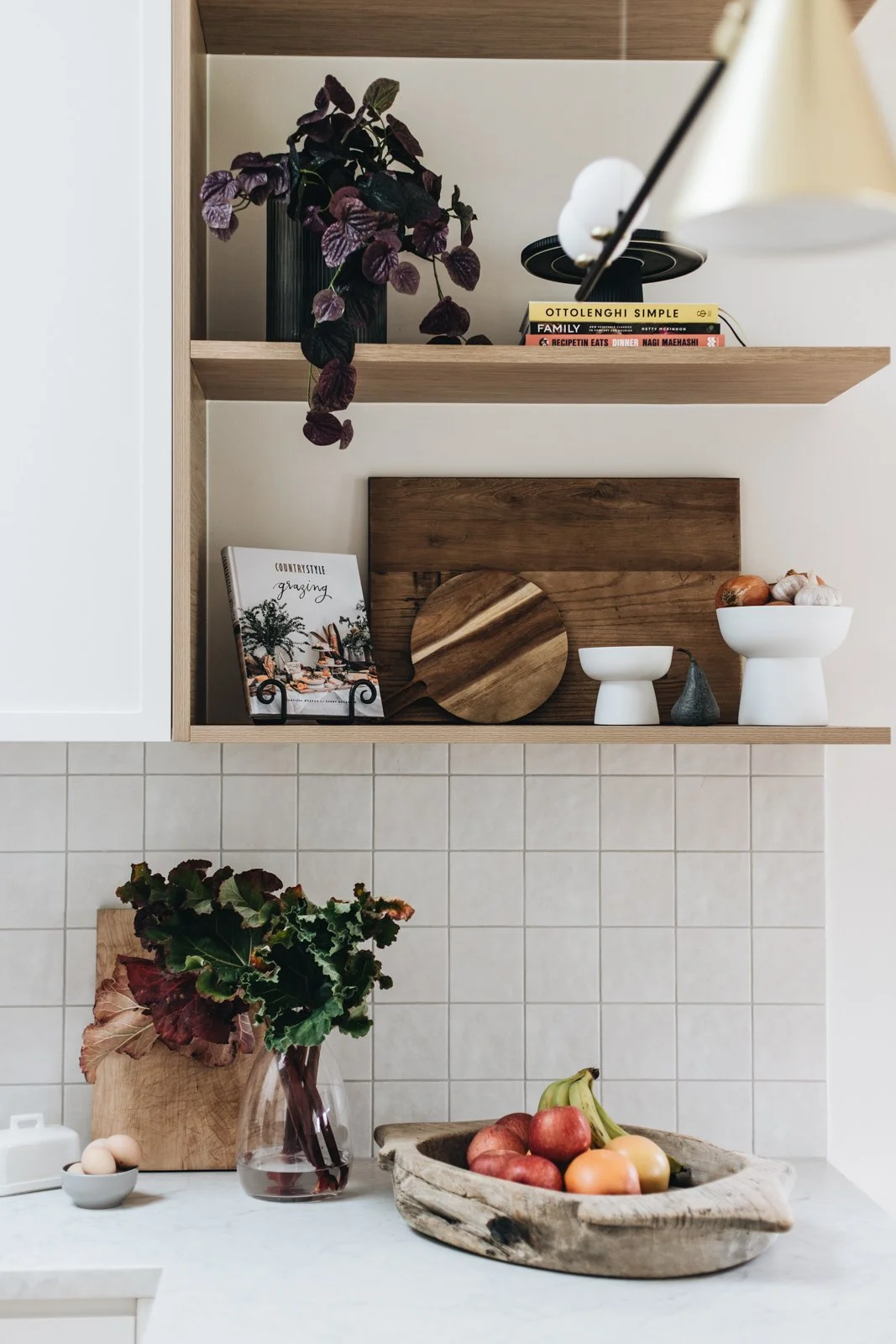 Kitchen shelf with potted plant, books, and decorative items; countertop with a vase of flowers, a wooden bowl of apples and onions, and a small bowl of eggs.