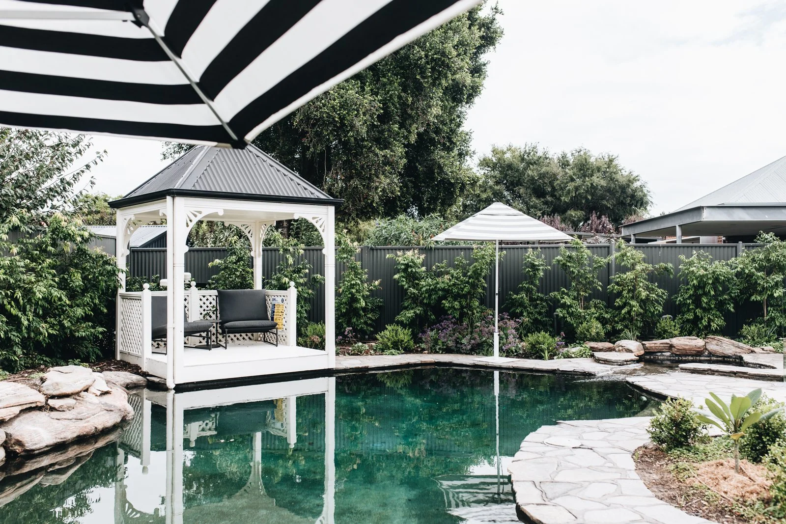 A backyard with a swimming pool, surrounded by greenery and a tall metal fence. There are two black and white striped umbrellas, a white gazebo with a bench inside, and a stone pathway beside the pool.