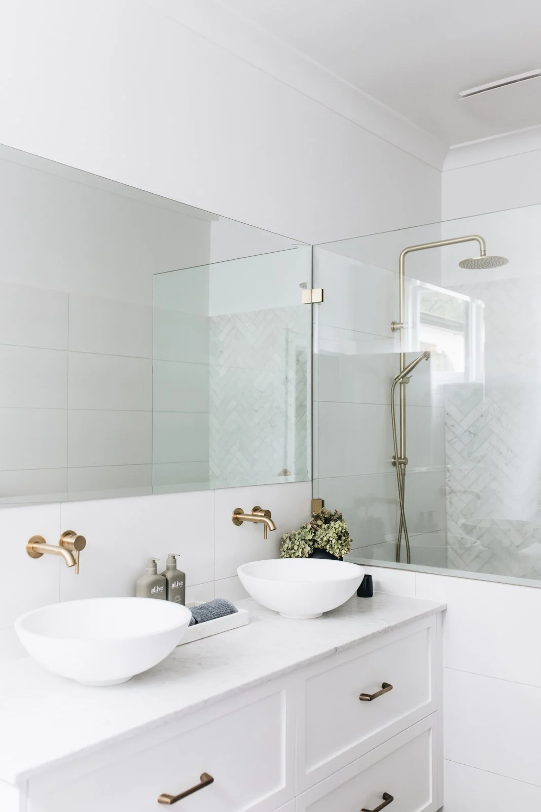 Modern white bathroom with double vessel sinks, brass faucets, large mirror, glass shower with a circular rainfall shower head, plants, black soap dispenser, and neatly arranged toiletries.