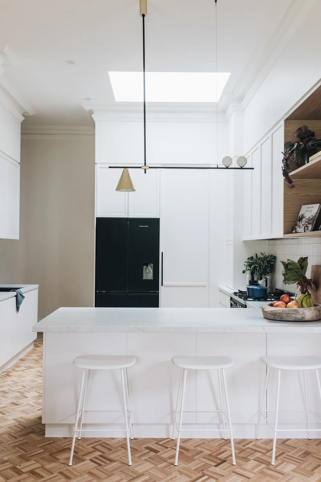Modern kitchen with white cabinets, black refrigerator, white marble island with three stools, wooden flooring, and open shelving with plants and books. Skylight overhead.