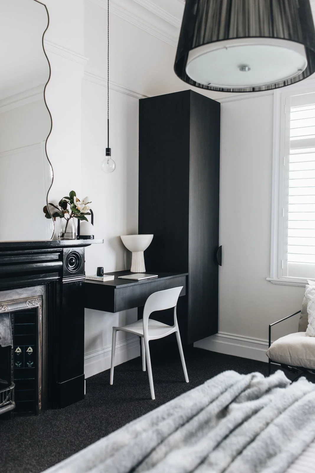 Modern bedroom with black and white decor, including a black cabinet, a white chair, a white vase, a black hanging light, a black and white mirror, and a bed with a textured blanket.