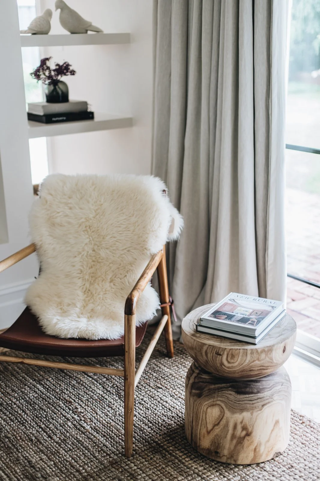 A cozy interior corner featuring a wooden armchair with a sheepskin throw, a marble side table with magazines, a window with curtains, and built-in shelves with decorative items.