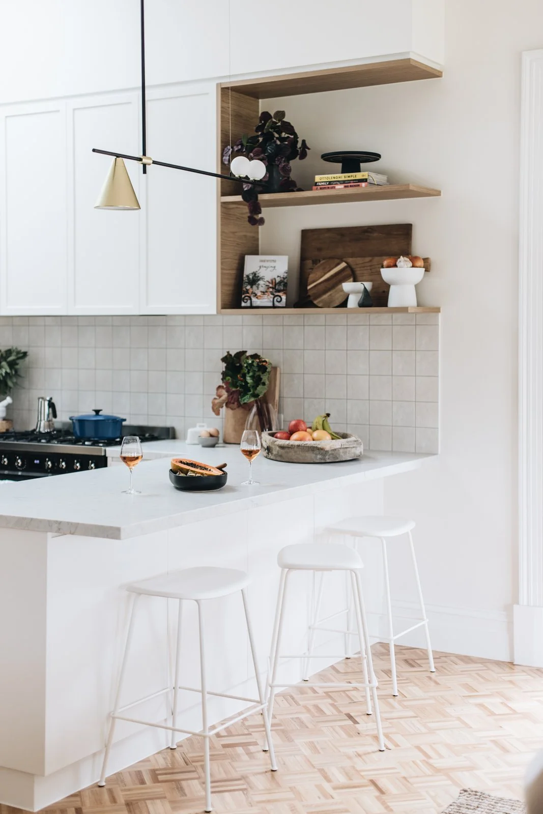 A modern kitchen with white cabinets and countertop, open wooden shelves holding decorative items and books, a stove with a blue pot, a bowl of papaya, two glasses of rosé wine, and bar stools at the counter.