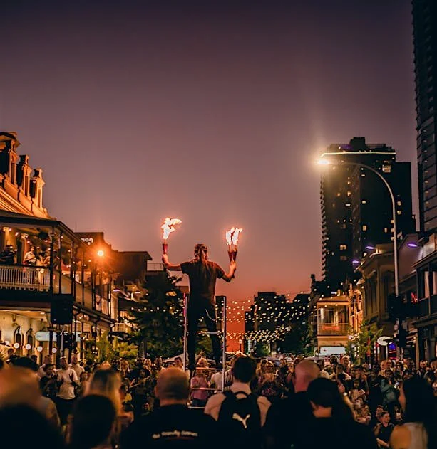 Street scene at dusk with a fire performer standing on stilts, holding torches, surrounded by a crowd of people, with buildings and string lights in the background.