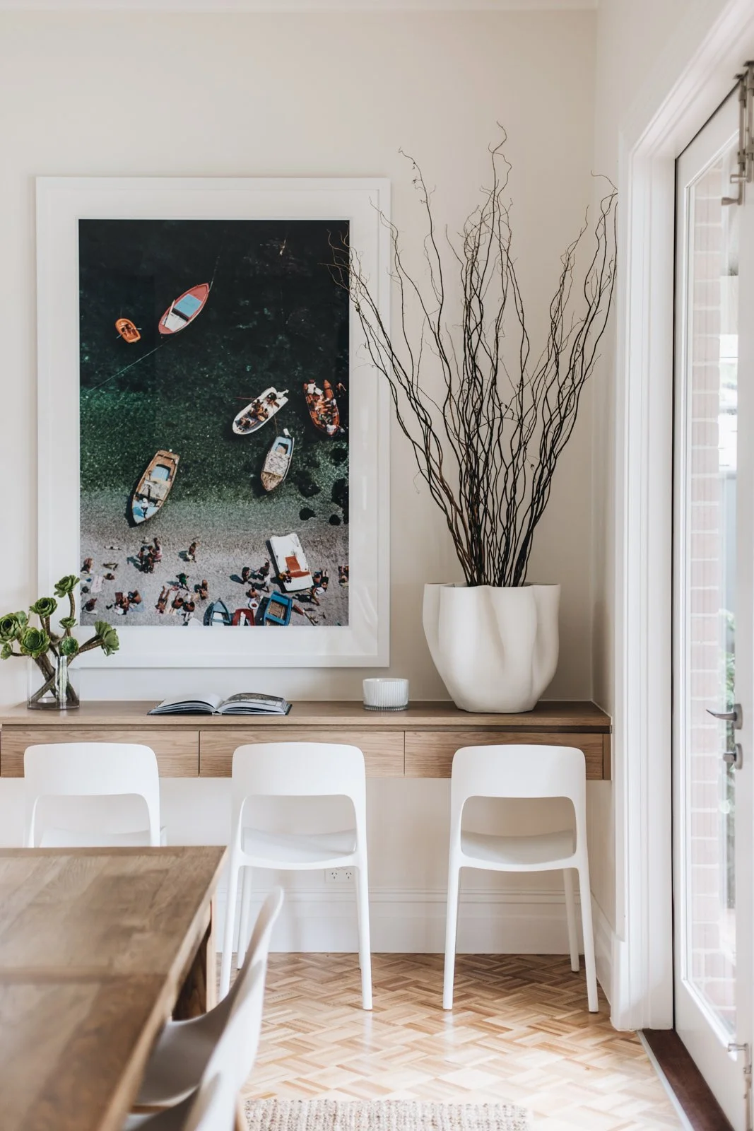 Living room corner with a large framed aerial photo of boats on water, a white vase with tall twig branches, a small vase with green flowers, an open book, and a small white container on a wooden sideboard, with three white chairs and a window nearby