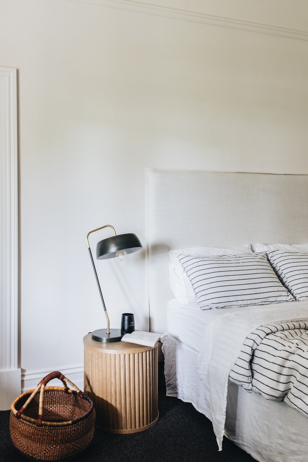 A bedroom with a white bed and striped pillows, black and gold modern table lamp on a round wooden side table, a book and a black cup, a woven basket on a dark carpeted floor, and a white wall with a ceiling molding.