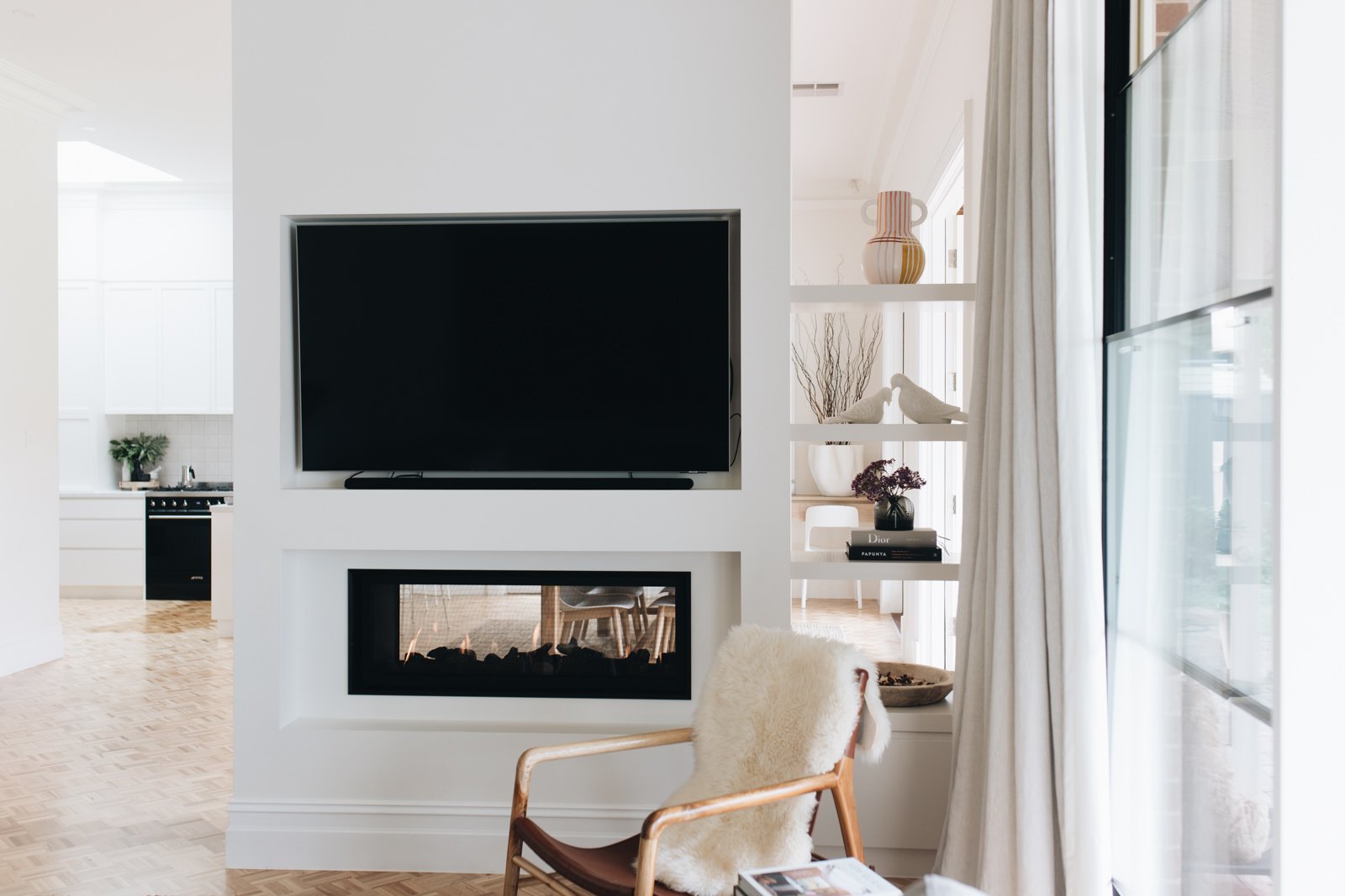 Living room with a wall-mounted flat-screen TV above a fireplace, a wooden armchair with a sheepskin throw, books, and decorative items on white shelves, next to large windows with curtains, and a view into a bright kitchen.