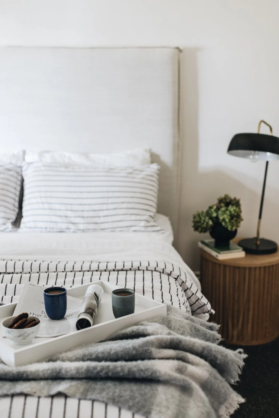 A cozy bedroom with a white bed featuring striped pillows and bedding, a tray with drinks and cookies on the bed, a wooden side table with a plant, books, and a black desk lamp, and a neutral wall color.