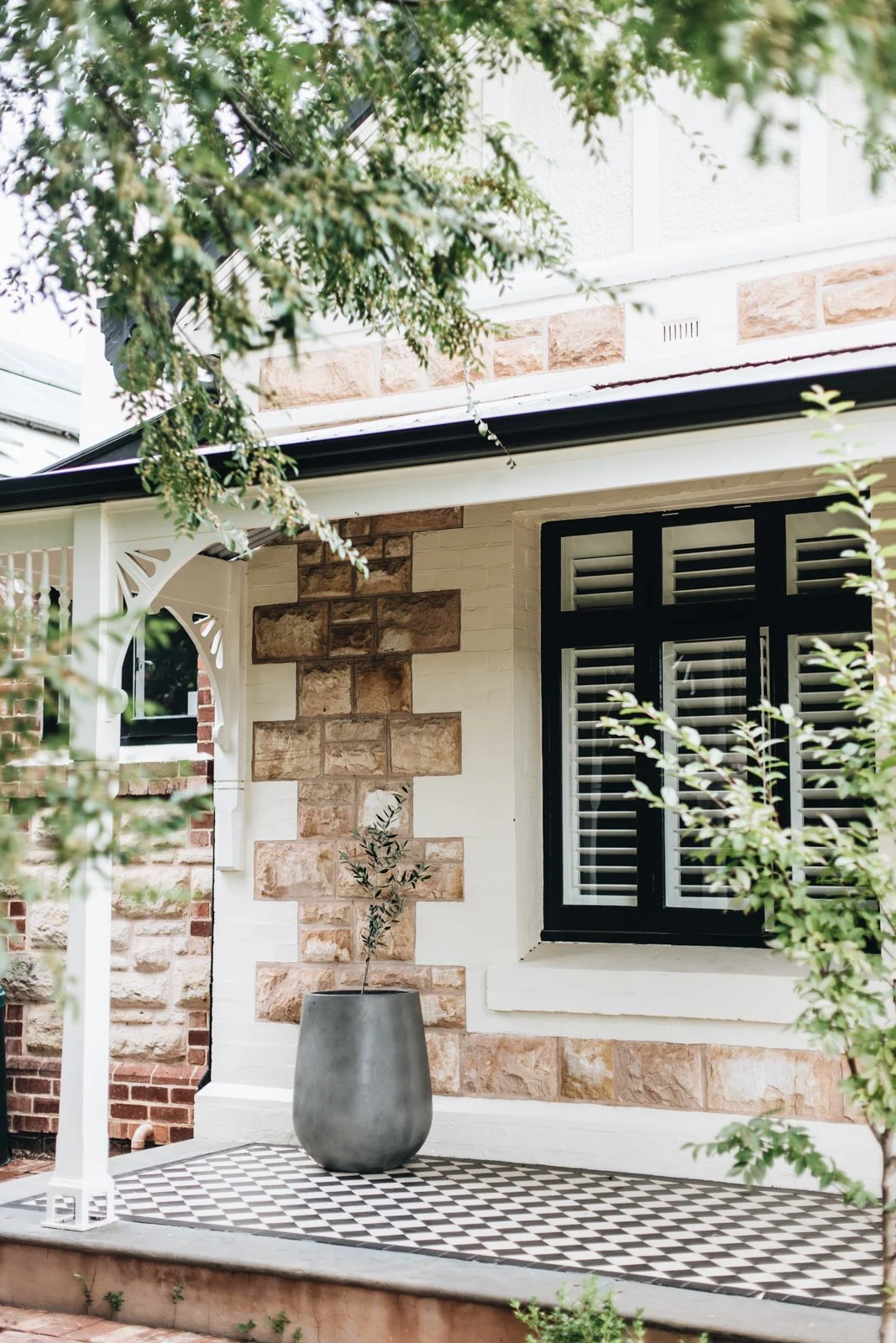 Close-up of a welcoming house front porch with black window shutters, a large gray planter with a small plant, and checkered black and white flooring, surrounded by green foliage and trees.