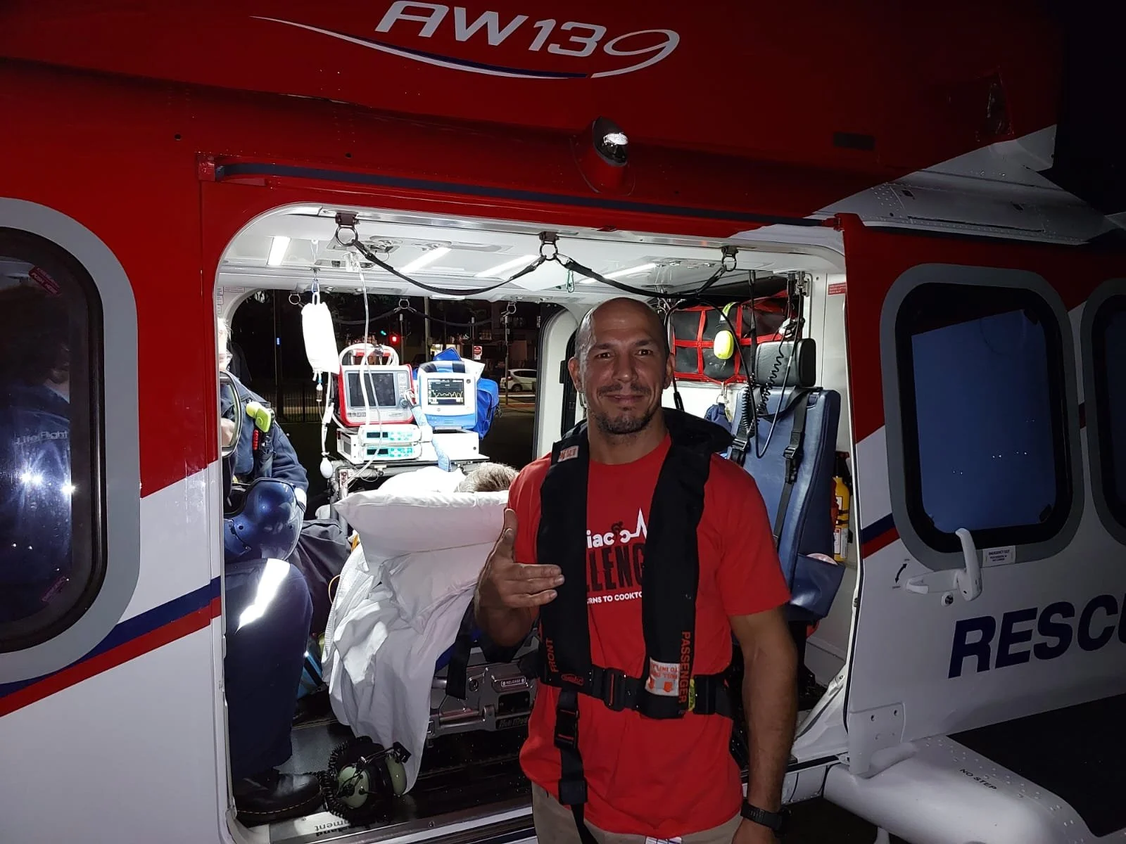 A man standing outside a rescue helicopter at night, making a hand gesture, with a paramedic and medical equipment visible inside the helicopter.