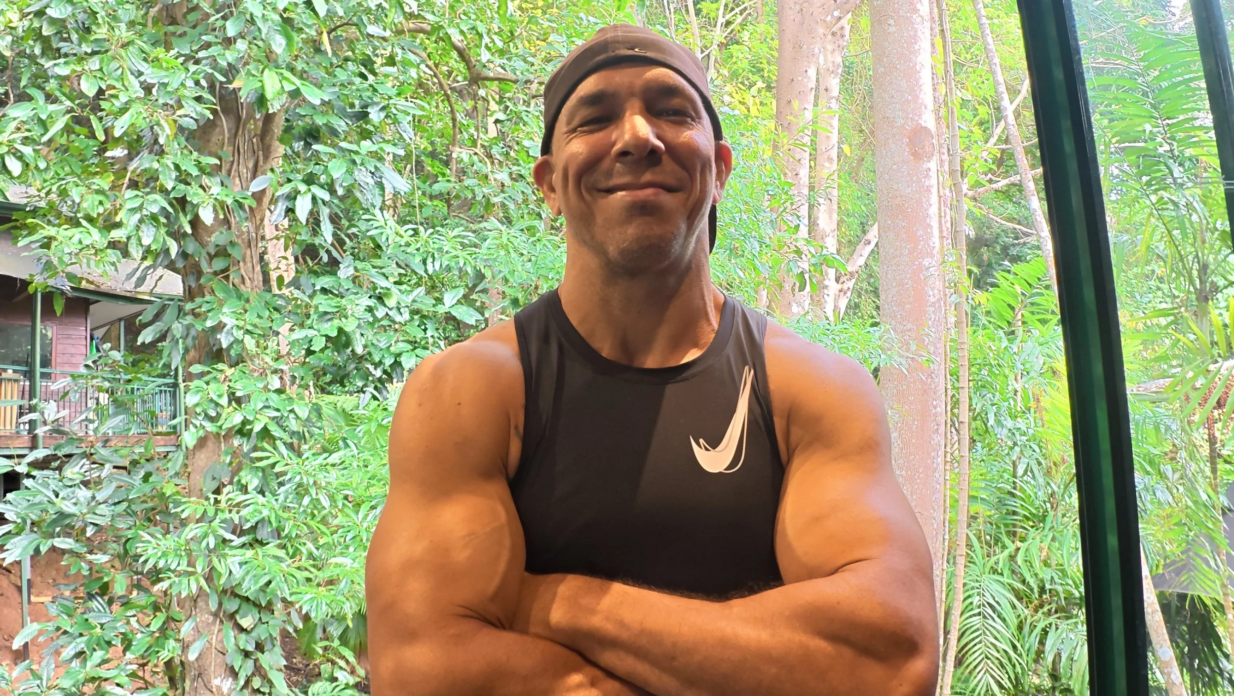 A man wearing a black Nike tank top and a black headband is standing outdoors with crossed arms, smiling, in front of lush green trees and foliage.