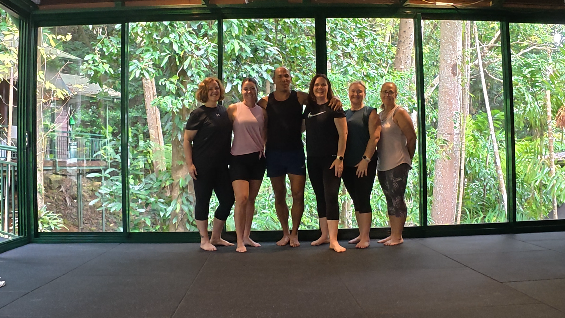 Group of six women standing barefoot inside with a view of lush green trees and foliage through large glass windows behind them