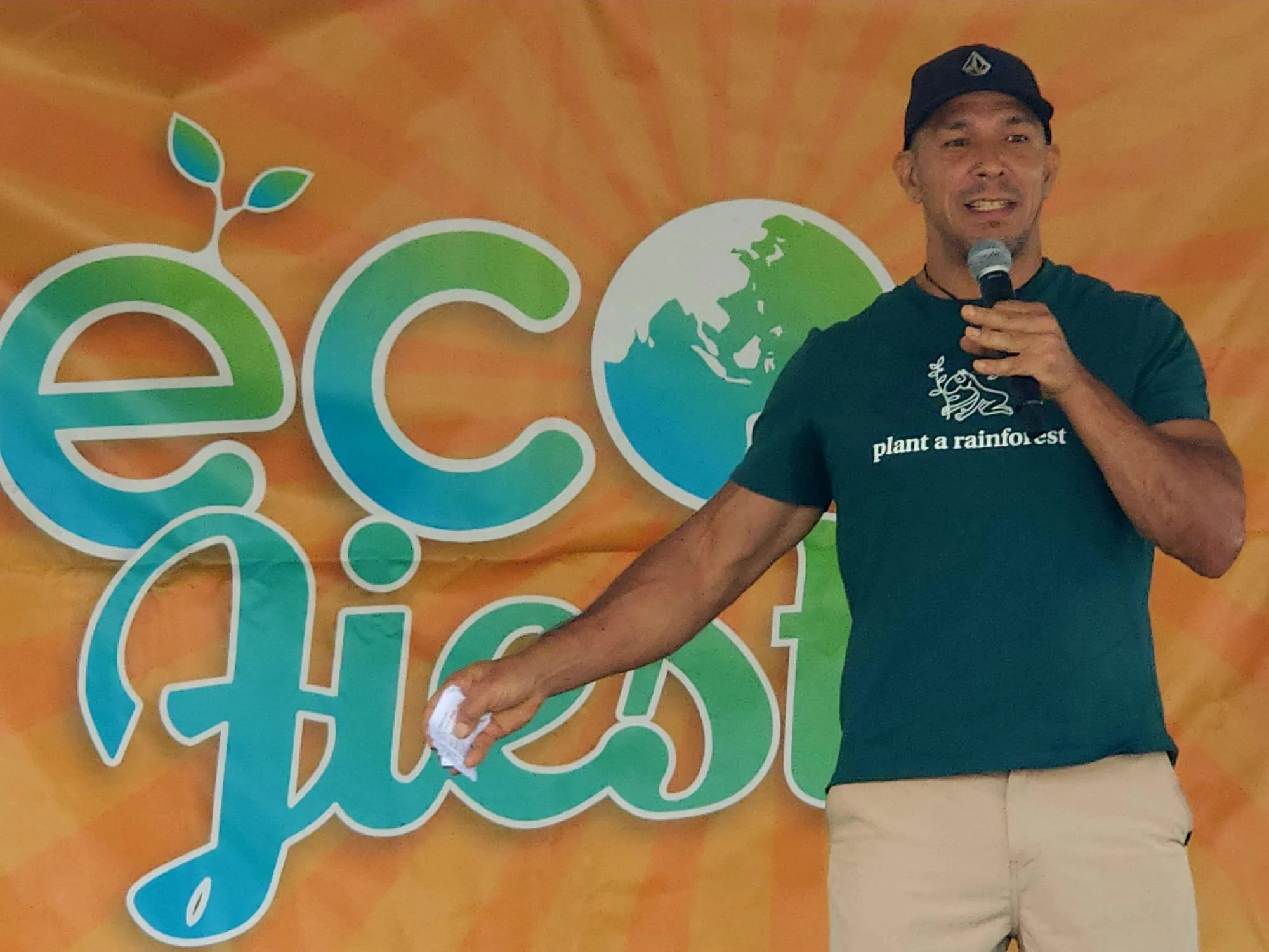 A man holding a microphone and a cloth stands in front of an orange background featuring a logo with a leaf, a globe, and a stylized tree. The man is wearing a black T-shirt with the words 'plant a rainforest' and a graphic of a frog.