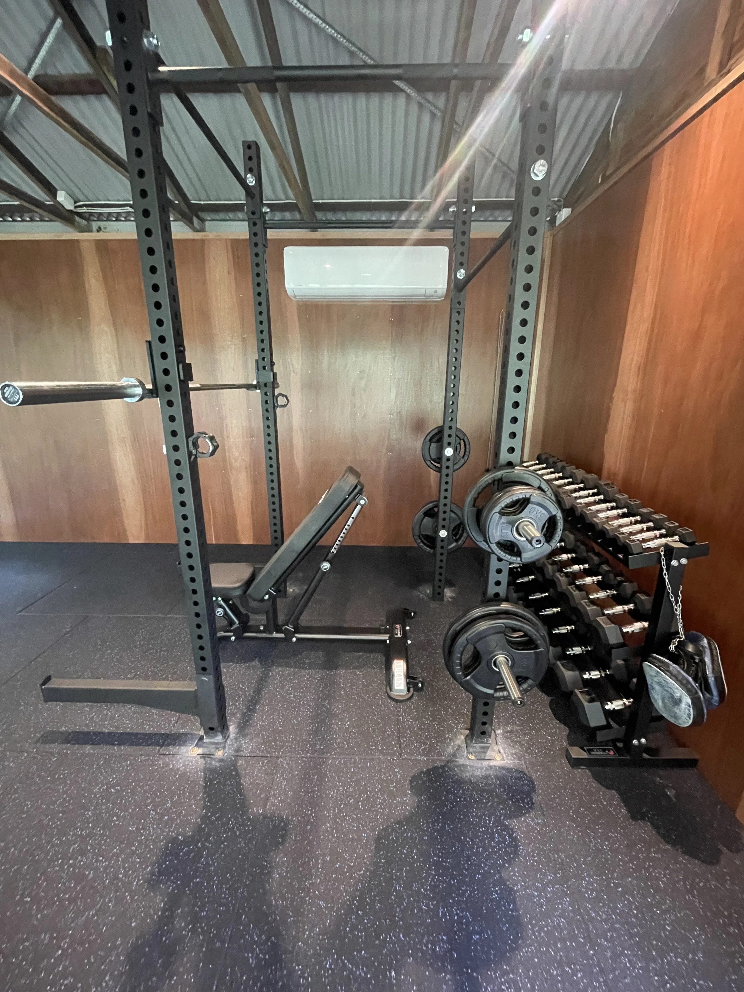Home gym with black metal squat rack, weight plates, dumbbells on rack, and pull-up bar, all on a black speckled rubber floor, with a wooden wall and an air conditioning unit in the background.