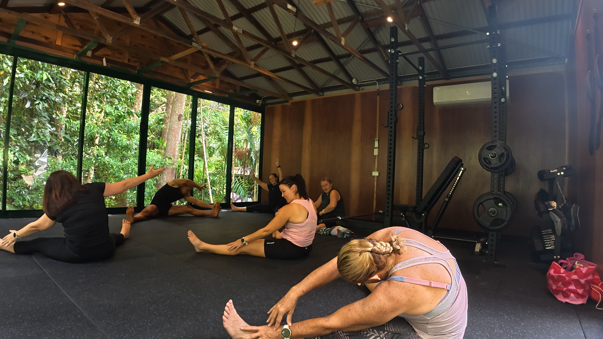 People practicing yoga in a studio with large windows showing a lush green outdoors, some in seated stretches and others in different yoga poses.
