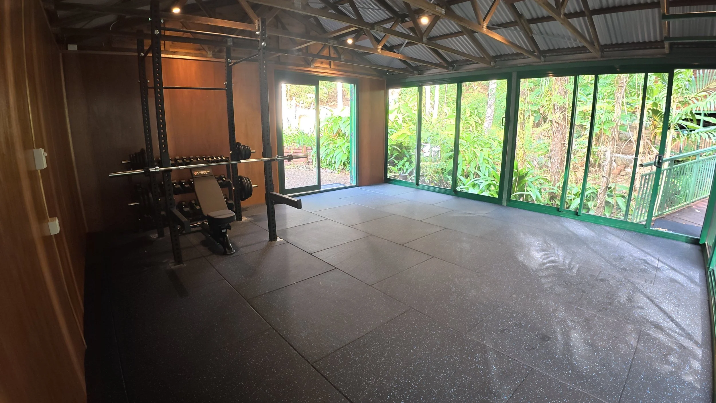 An indoor gym space with black rubber flooring, a large black metal squat rack, a weight bench, and view of lush green trees outside through large sliding glass windows.