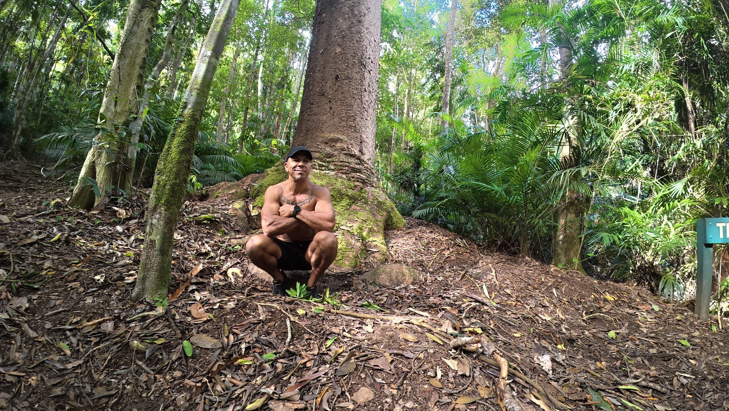 A shirtless man crouching in front of a large tree in a dense green jungle, smiling and crossing his arms.