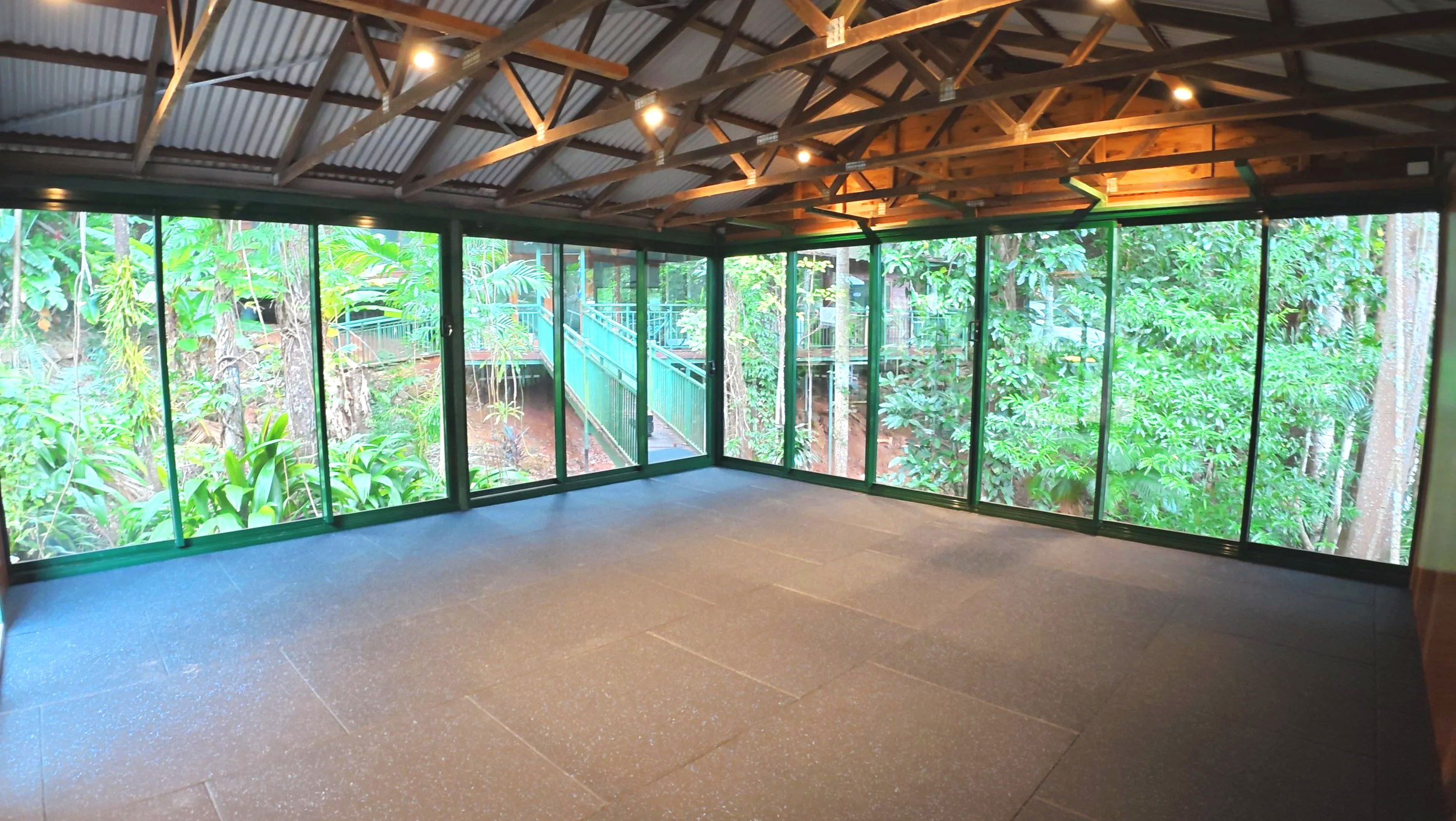 View from inside a building with large glass windows overlooking lush green tropical trees and foliage, with a bridge visible outside.