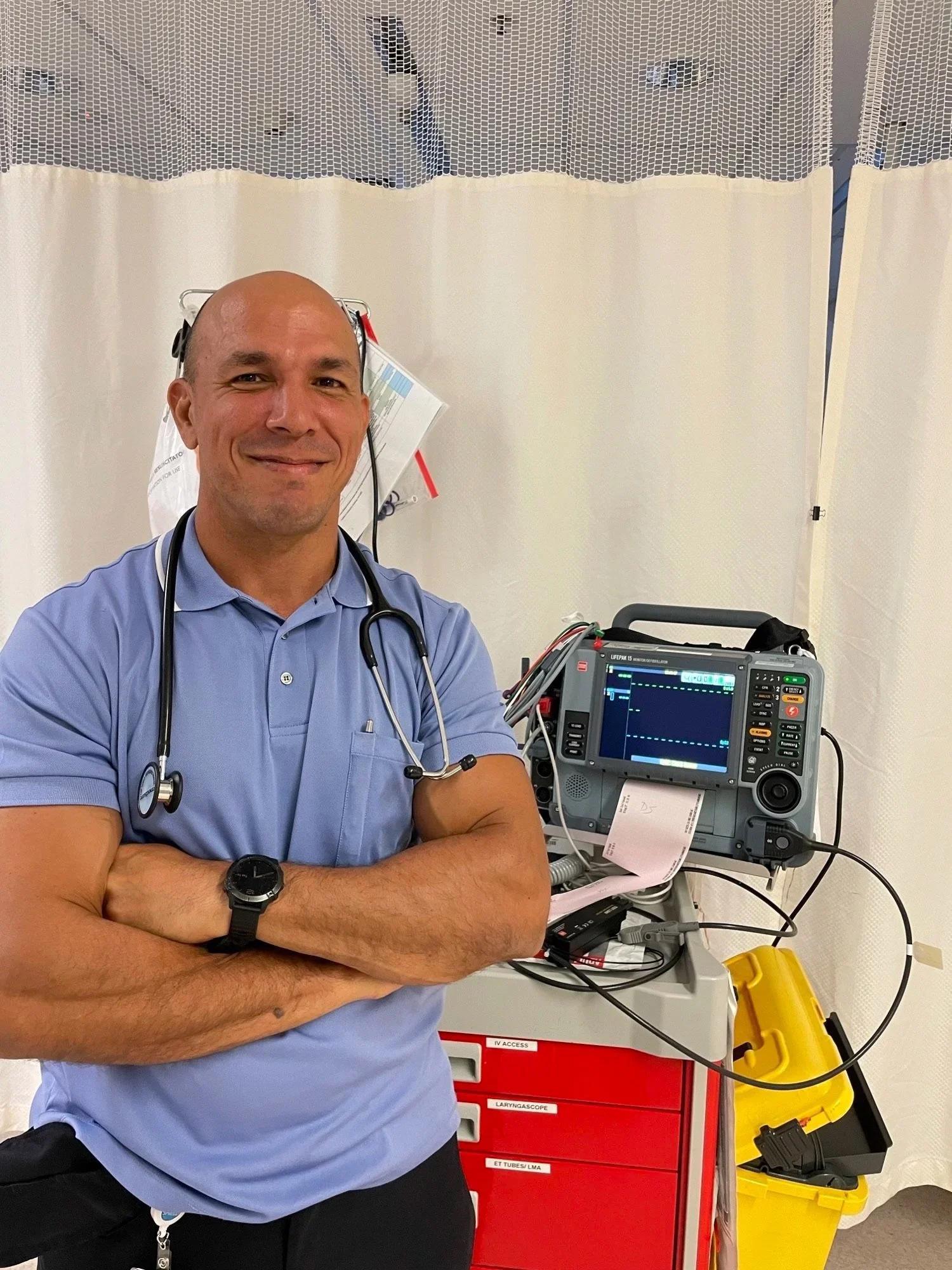 A male healthcare worker with a stethoscope around his neck, smiling at the camera in a medical setting, standing beside medical equipment including a monitor and a red medical storage cabinet.