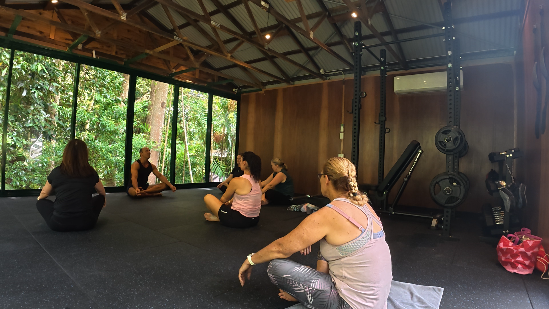 Group of women attending a yoga or meditation class in a rustic studio with large windows overlooking greenery.