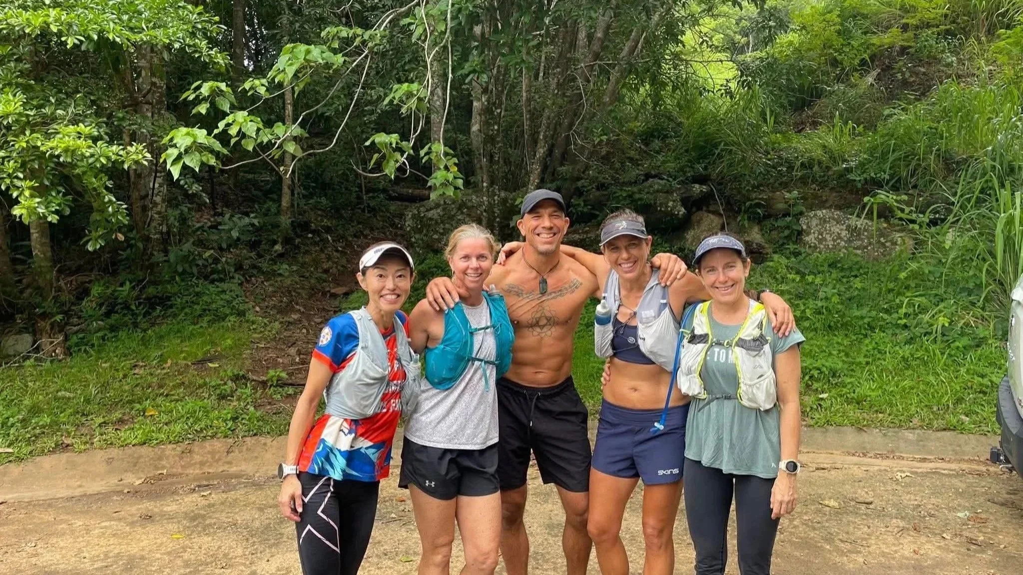 Group of five smiling trail runners posing together in a green forest. Four women and one shirtless man, all wearing athletic gear and hydration packs.