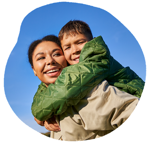 A woman and a young boy smiling and hugging outdoors under a clear blue sky.