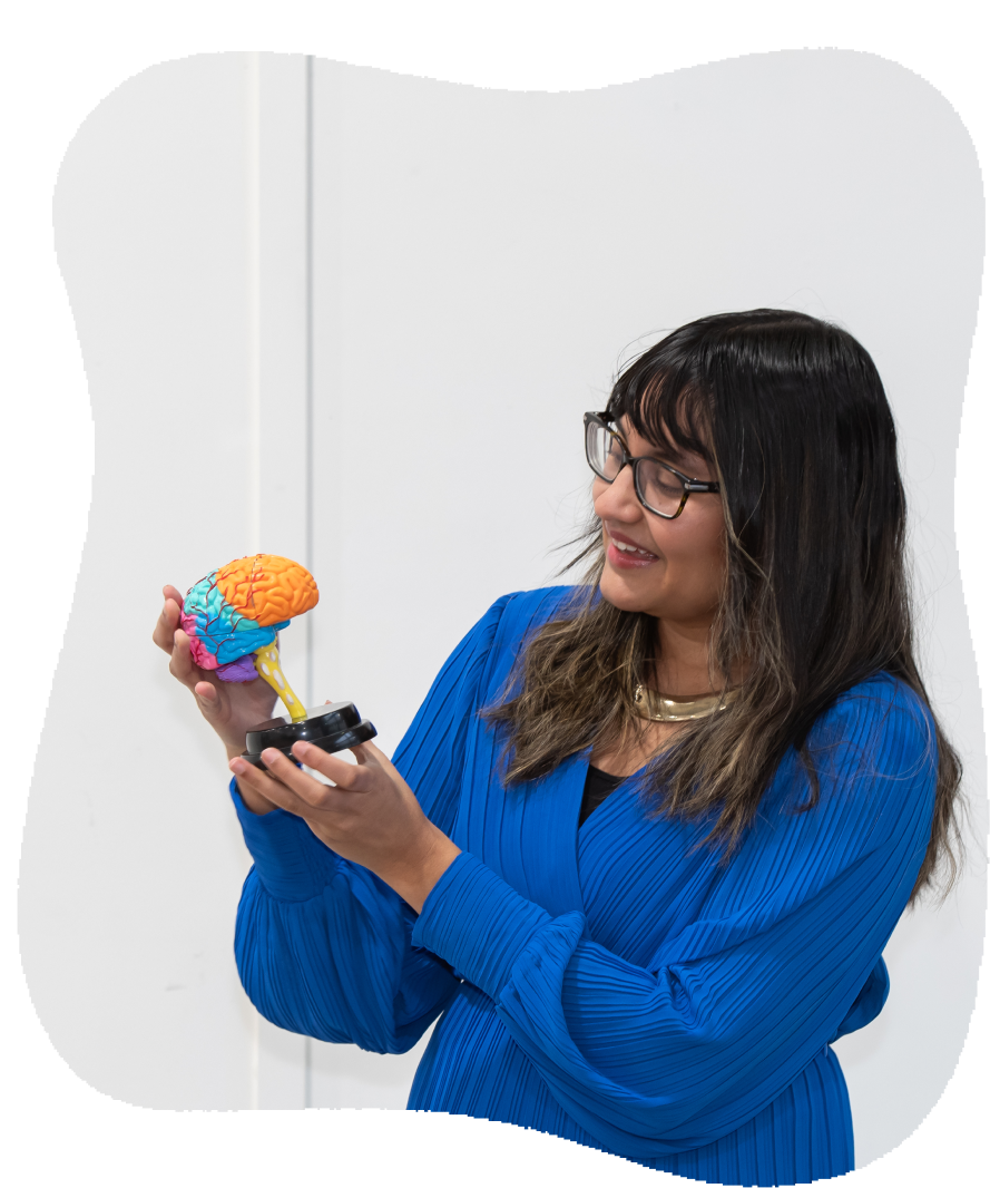 A woman with dark hair, glasses, and a blue dress holds a colorful model of a human brain.