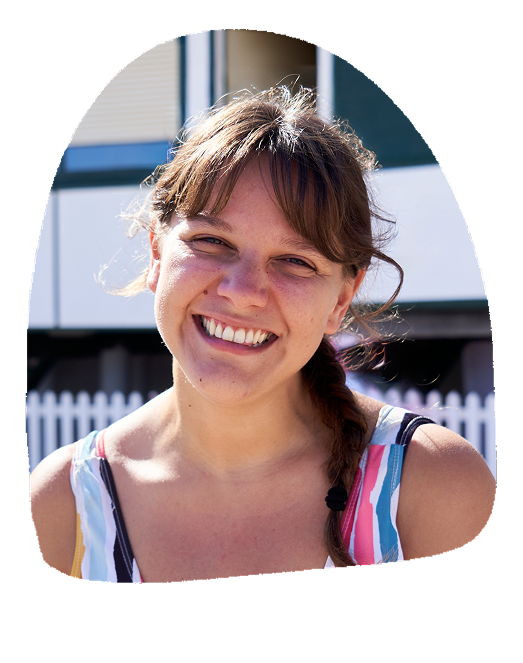 A young woman with brown hair, styled in loose waves with a side braid, smiling outdoors on a sunny day, wearing a striped tank top.