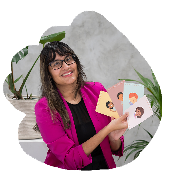 A woman wearing glasses and a bright pink blazer smiling and holding up flashcards with diverse children illustrations. There are potted plants in the background.