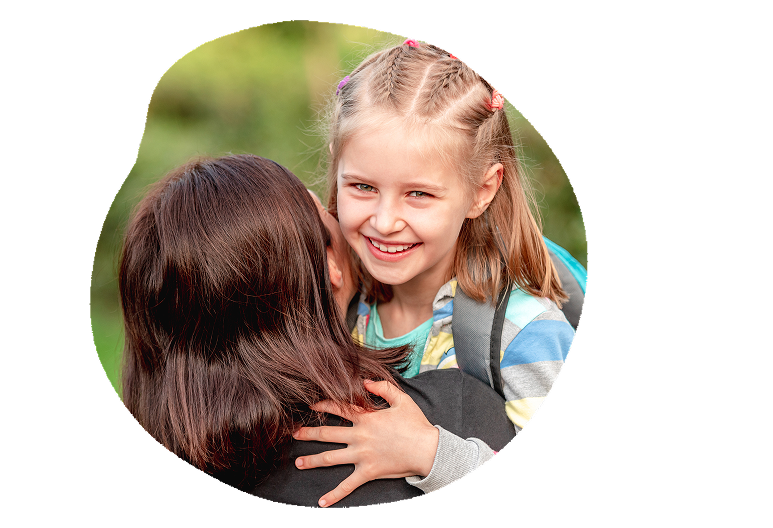 A young girl with blonde hair smiling as she hugs another person with dark brown hair outdoors.