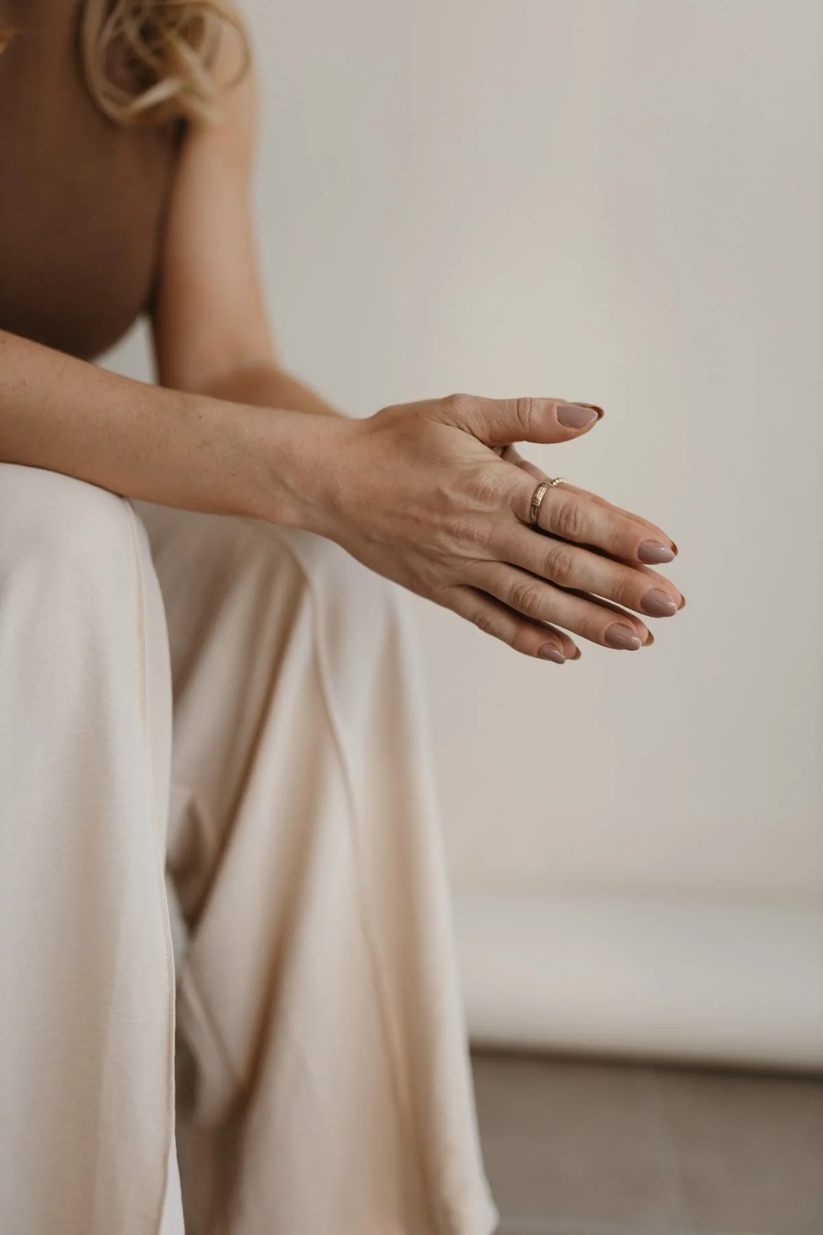 Close-up of a woman's hand resting on her knee, wearing a gold ring, with part of her arm and beige clothing visible, against a neutral background.