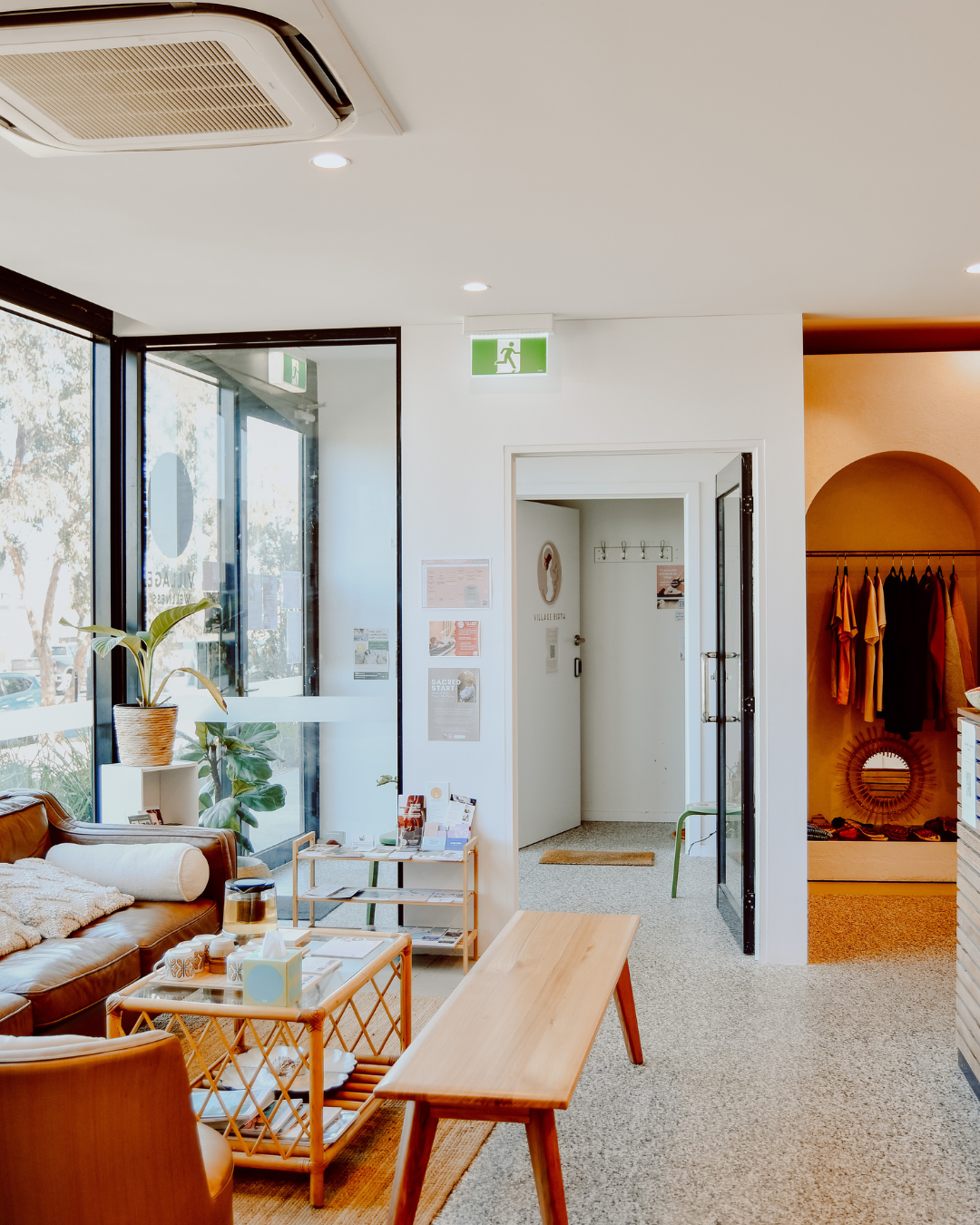 Interior of a waiting area or lobby with a brown leather sofa, a wooden coffee table, potted plants, and large windows allowing natural light. There is an exit sign above a doorway, and a small room with a coat rack and mirror visible in the background.