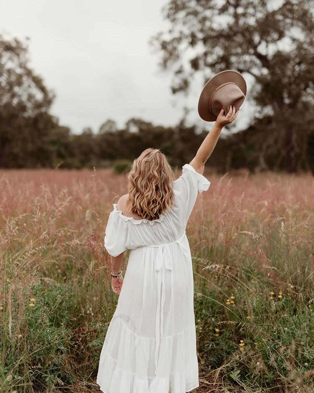 A woman with wavy blonde hair wearing a white dress and a bracelet, standing in a field of tall grass and flowers, holding a brown hat in the air with her right hand, with trees and an overcast sky in the background.