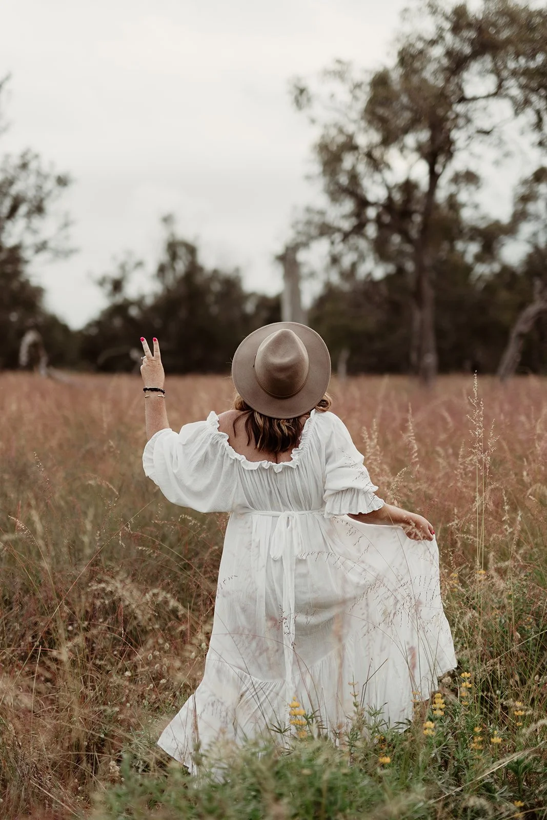 A woman in a white dress and wide-brimmed hat standing in a field of tall grasses and wildflowers, facing away with one arm raised, making a peace sign against a background of trees and cloudy sky.