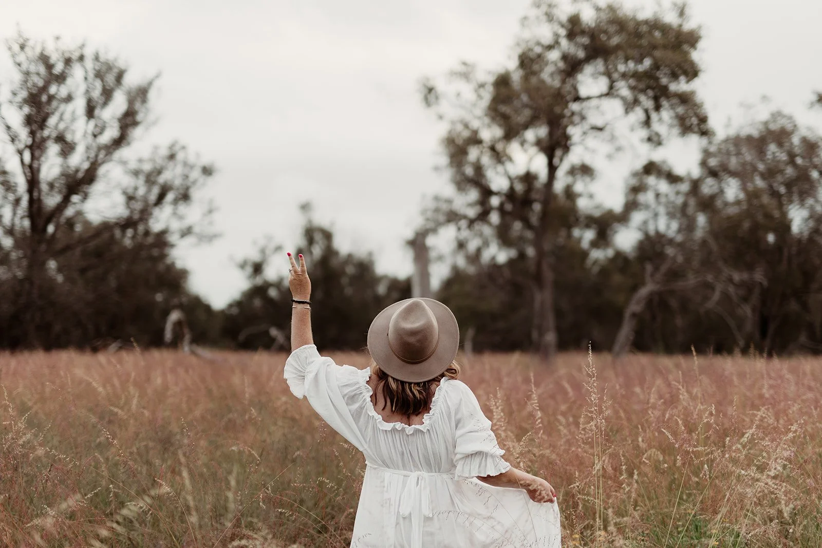 A woman in a white dress and beige wide-brim hat standing in a field of tall grass, with her back to the camera, raising her left hand with two fingers up. Trees are visible in the background under an overcast sky.
