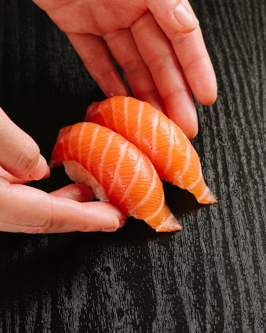 Two pieces of raw salmon nigiri sushi on a black wooden surface, held gently by a person's fingers.
