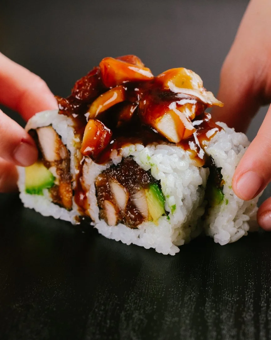 Close-up of a hand holding a sushi roll filled with avocado, crab or fish, and rice, topped with eel sauce and chopped cooked eel.