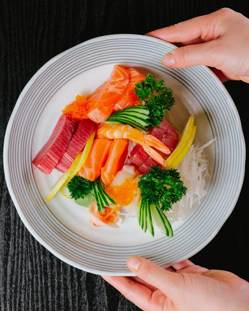 Platter of assorted sashimi including salmon, tuna, white fish, and shrimp, garnished with cucumber slices, parsley, grated radish, and orange fish roe, served on a bed of shredded daikon radish.
