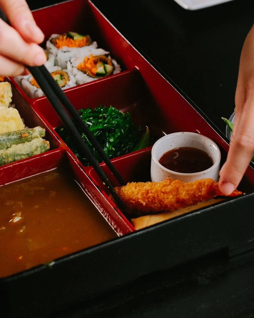 A bento box with sushi rolls, tempura vegetables, seaweed salad, curry, and a side of dipping sauce with a piece of fried fish being dipped.