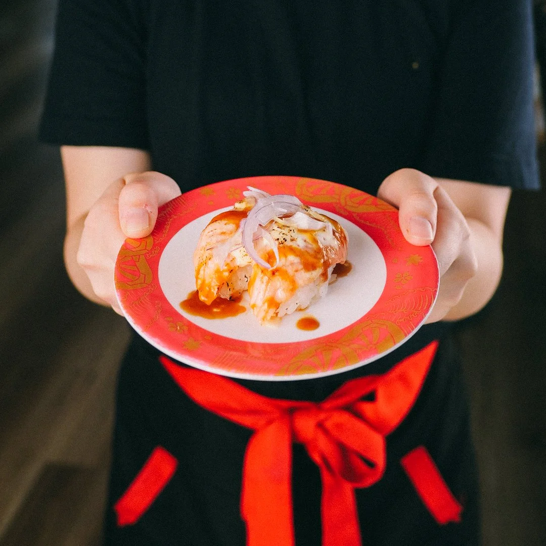Person holding a small plate with sushi topped with sauce and onions, wearing a black shirt and an apron with a red tie.