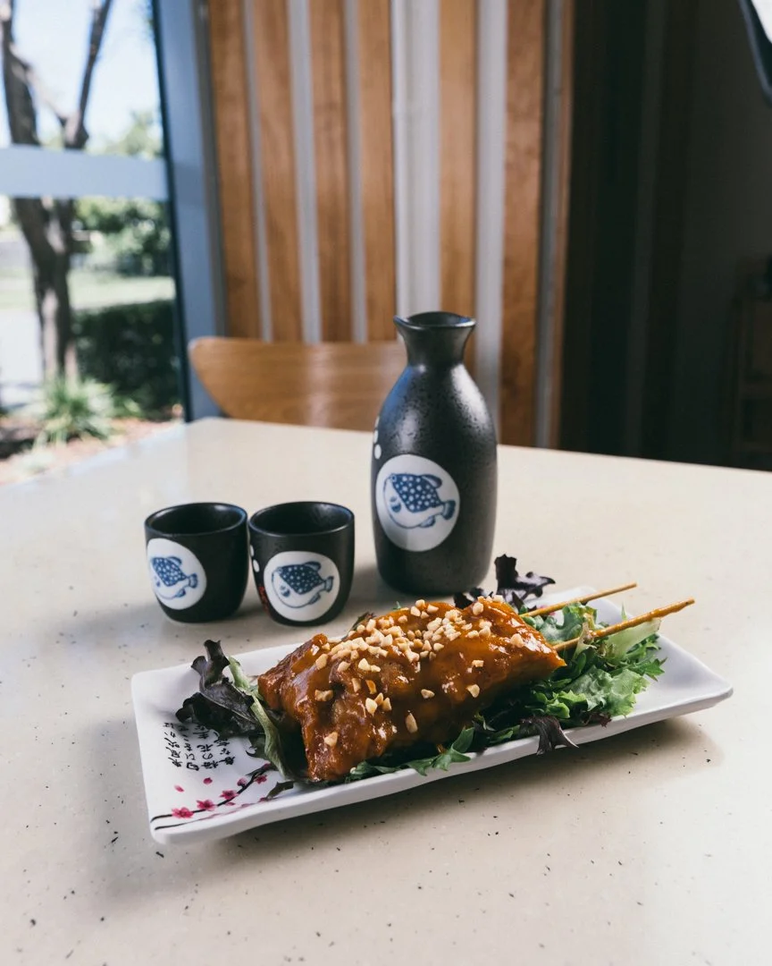 A plate of glazed meat skewers garnished with chopped nuts, lettuce, and greenery, with a small black and white sake set and a matching sake bottle on a white table, with a wooden wall and window in the background.