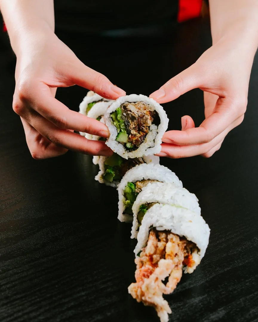 Person holding a piece of sushi with cucumber and tempura shrimp, with four other sushi pieces arranged in a line on a black surface.