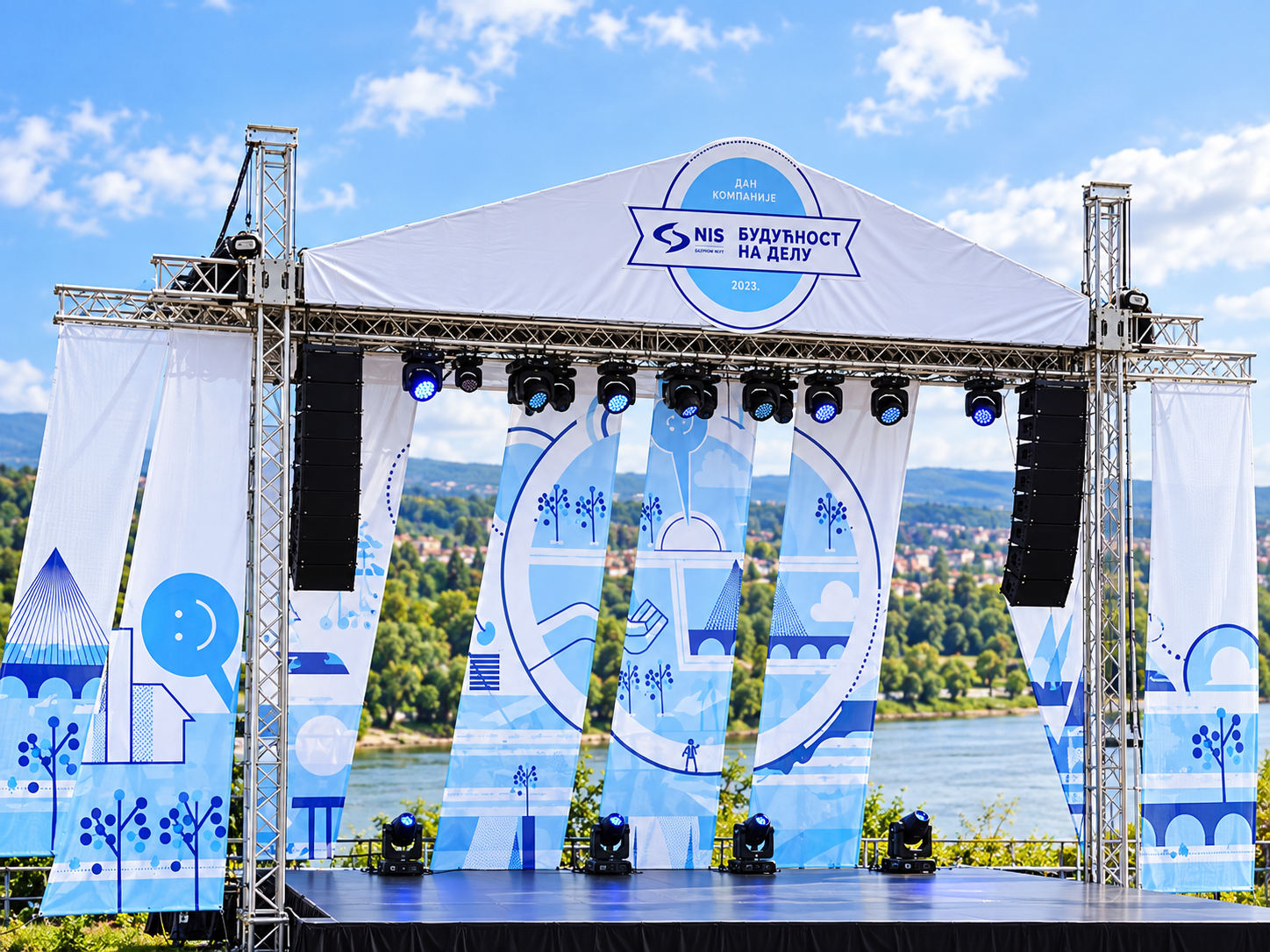 An outdoor stage with a white canopy and blue themed banners and panels, set against a backdrop of a river and green hills under a partly cloudy sky.