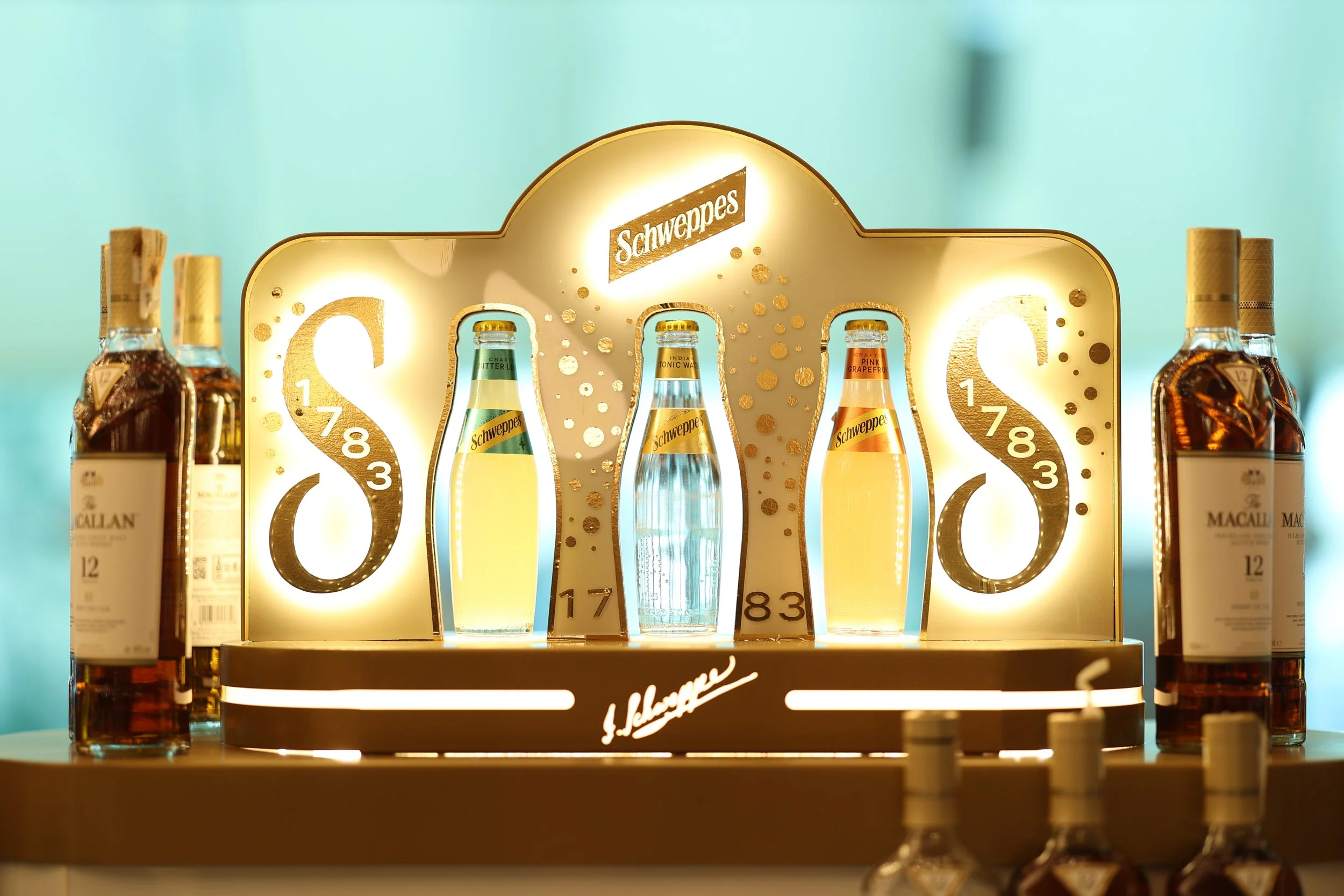 A display of Schweppes tonic water bottles with a backlit sign featuring the Schweppes logo and gold-colored decorative elements.
