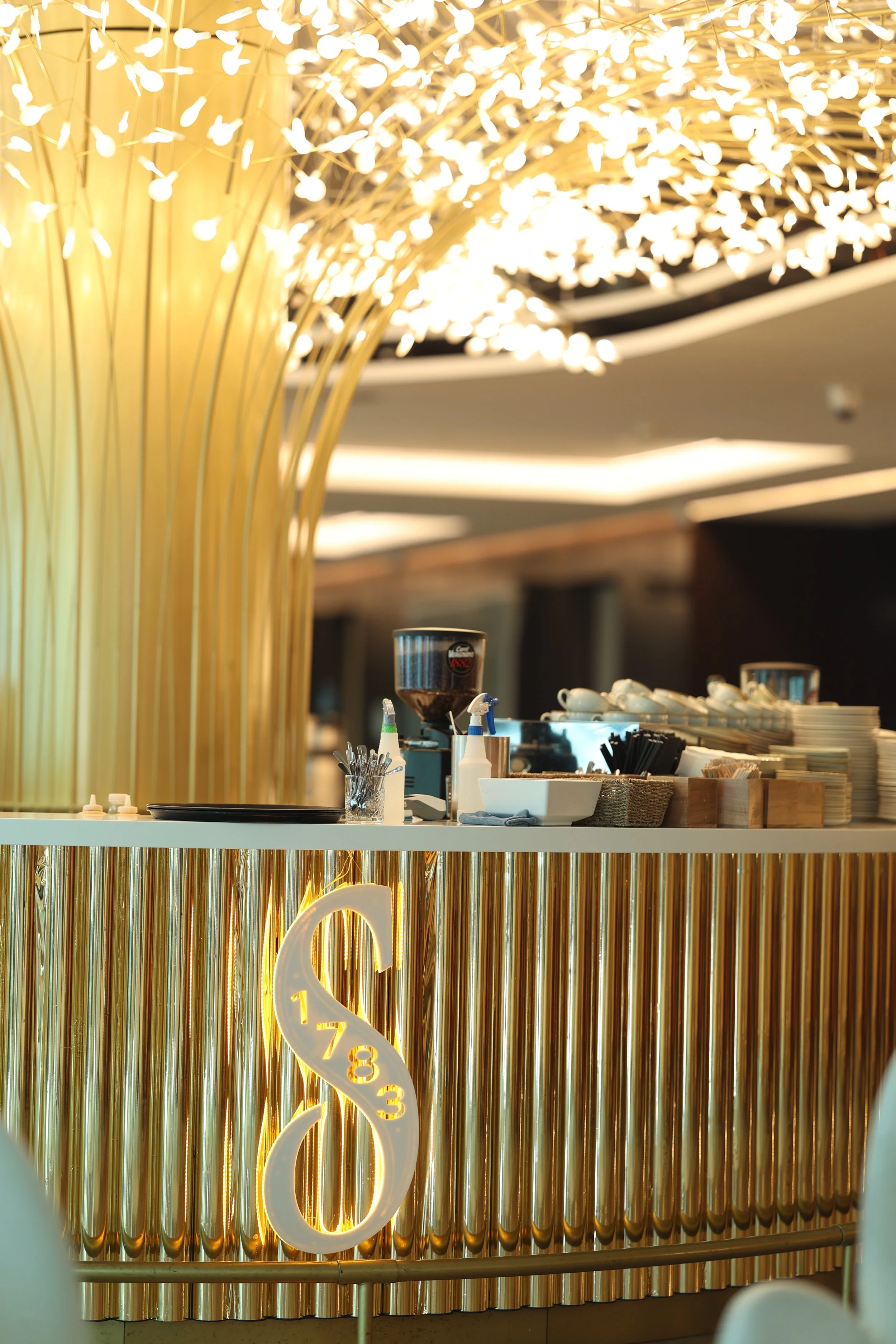 Close-up of a modern cafe counter with a gold ribbed front panel, illuminated with a large, decorative illuminated ampersand (&) sign, and a background of warm lighting and shelves with cups and kitchen items.