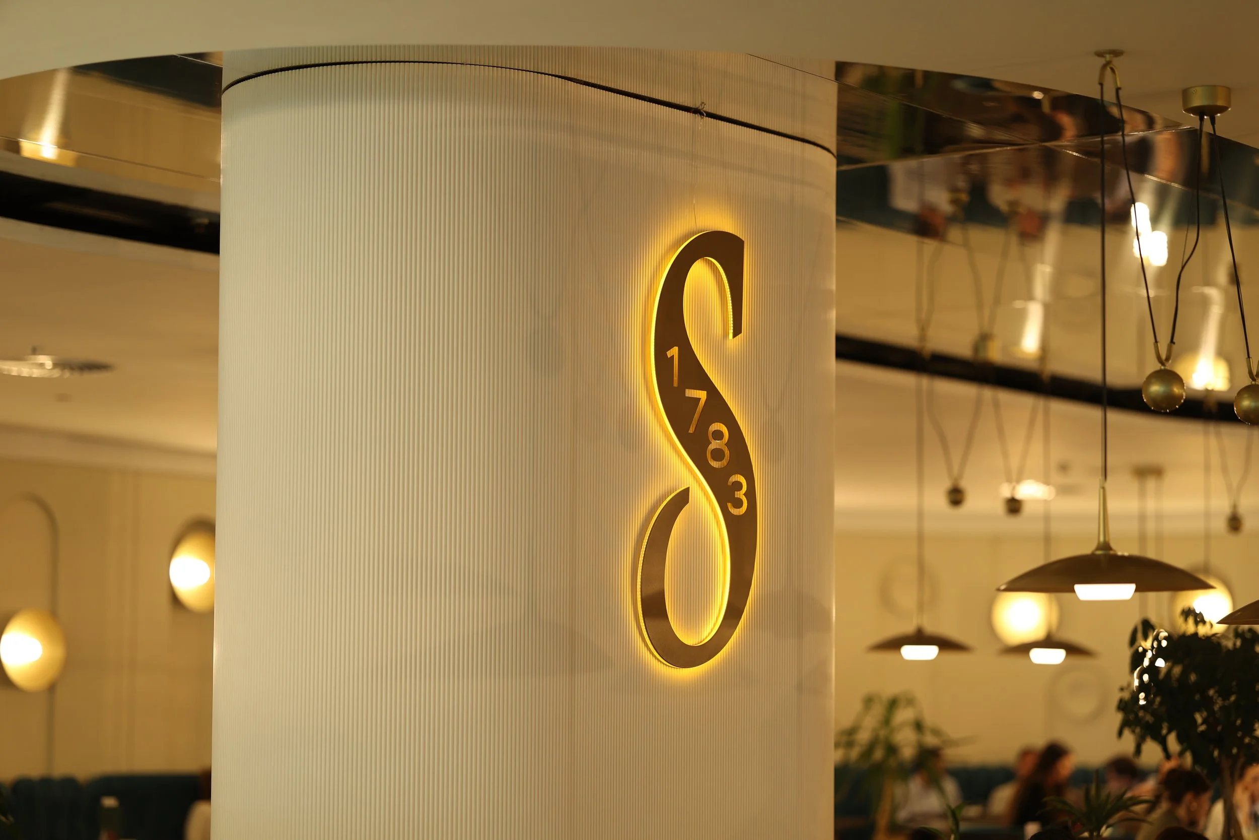 A column with a round logo with a yellow outline and numbers 1, 7, 8, 3, arranged in a pattern. The background has warm lighting and hanging fixtures.