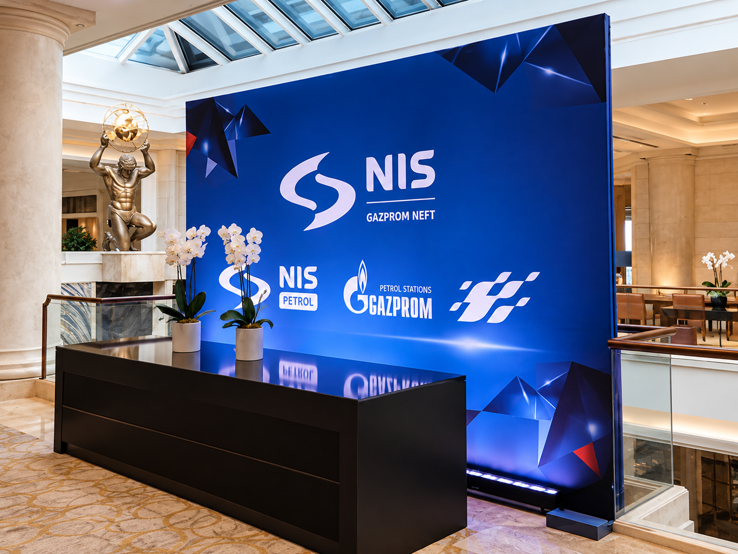 A large blue display board featuring the Gazprom logo and related branding, positioned in a spacious, well-lit interior with marble flooring, a statue of a man holding a globe, and potted white orchids on a black table in front of the display.